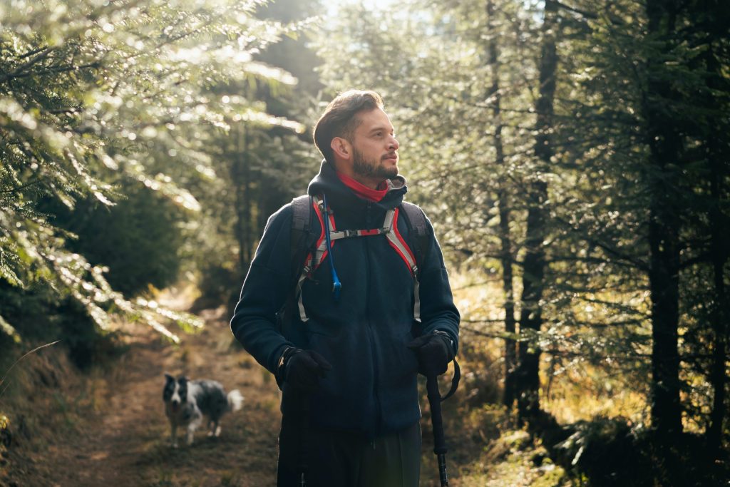 How Long Does Trauma Therapy Take? 4 Hiker in a dark jacket with a backpack on a sunlit forest trail, a dog visible in the background.